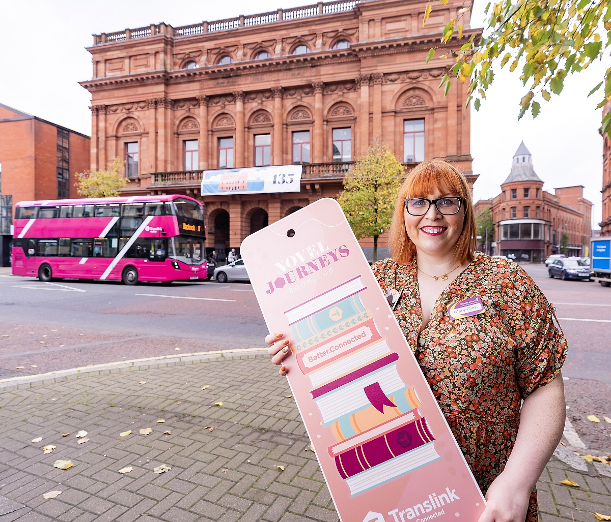 Librarian outside Belfast Central Library holding Translink's Novel Journeys sign and Translink bus
