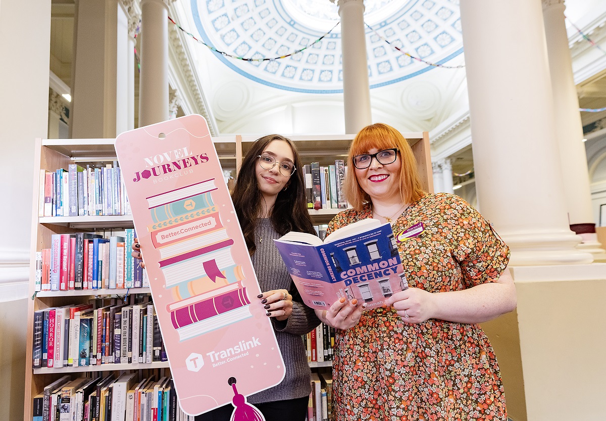 Two librarians inside Belfast Central Library holding Translink's Novel Journeys sign
